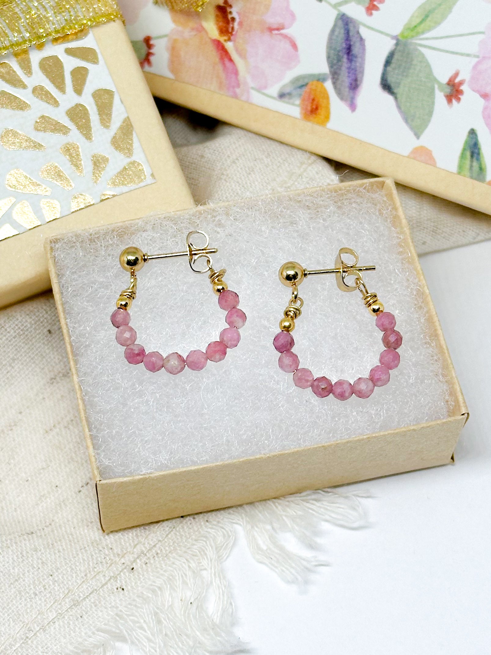 A pair of tourmaline birthstone hoop earrings presented in a beige box with a white interior, adorned with small pink gemstones.